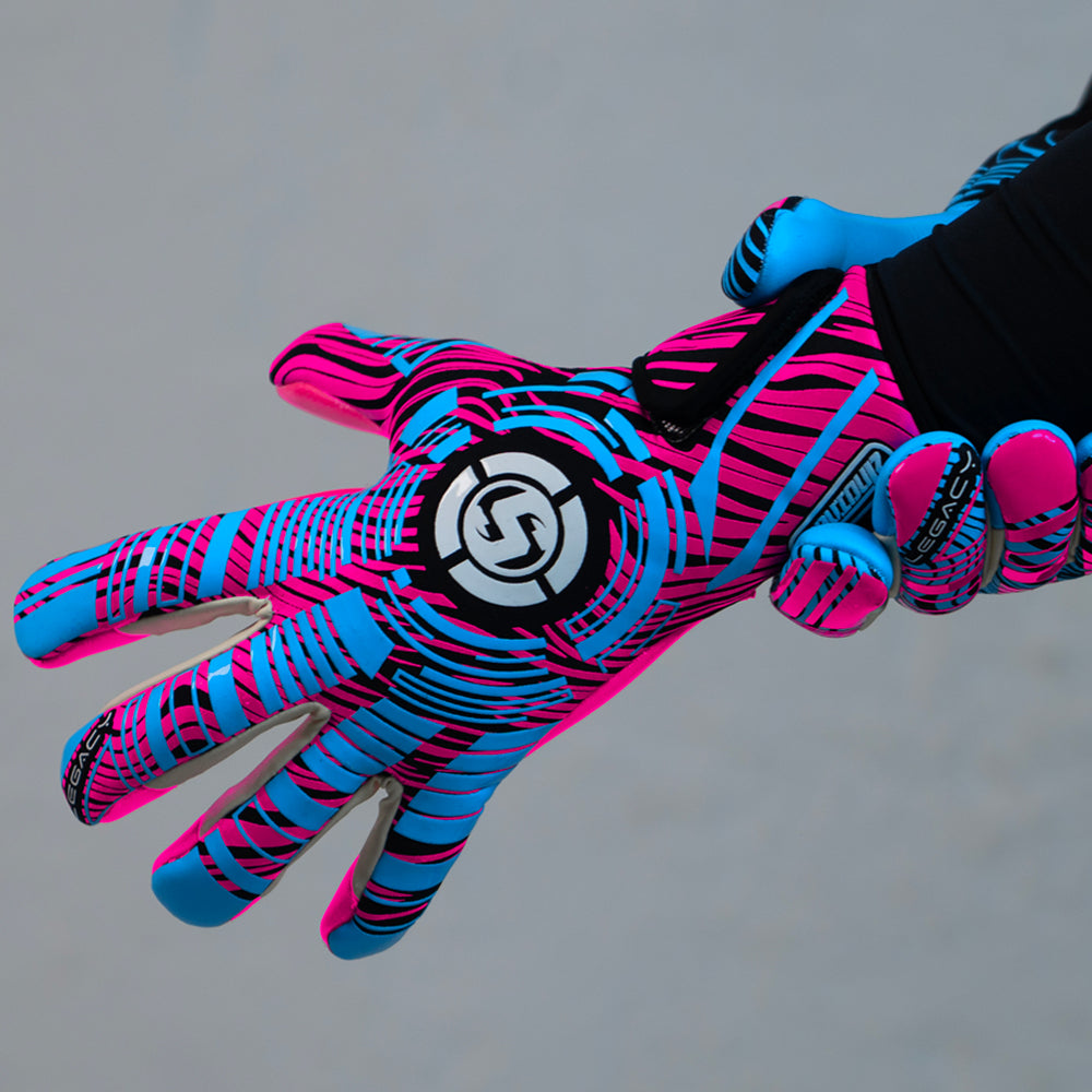 A close-up of a goalkeeper glove worn on a hand, featuring a vibrant neon pink and blue pattern with black accents and a circular logo on the back of the hand.