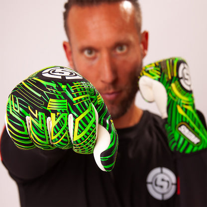 A man wearing green and black goalkeeper gloves with white accents, pointing one glove toward the camera against a plain light background.