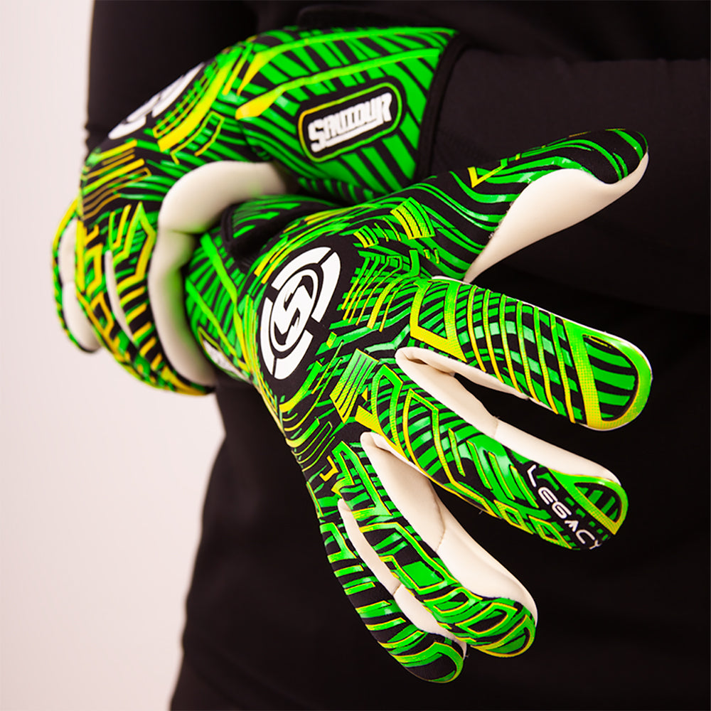 A close-up of a person wearing green and black patterned goalkeeper gloves with white contact palm grip, shown against a black shirt and light background.